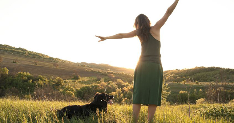 A image of a young women and a black dog facing to sun set