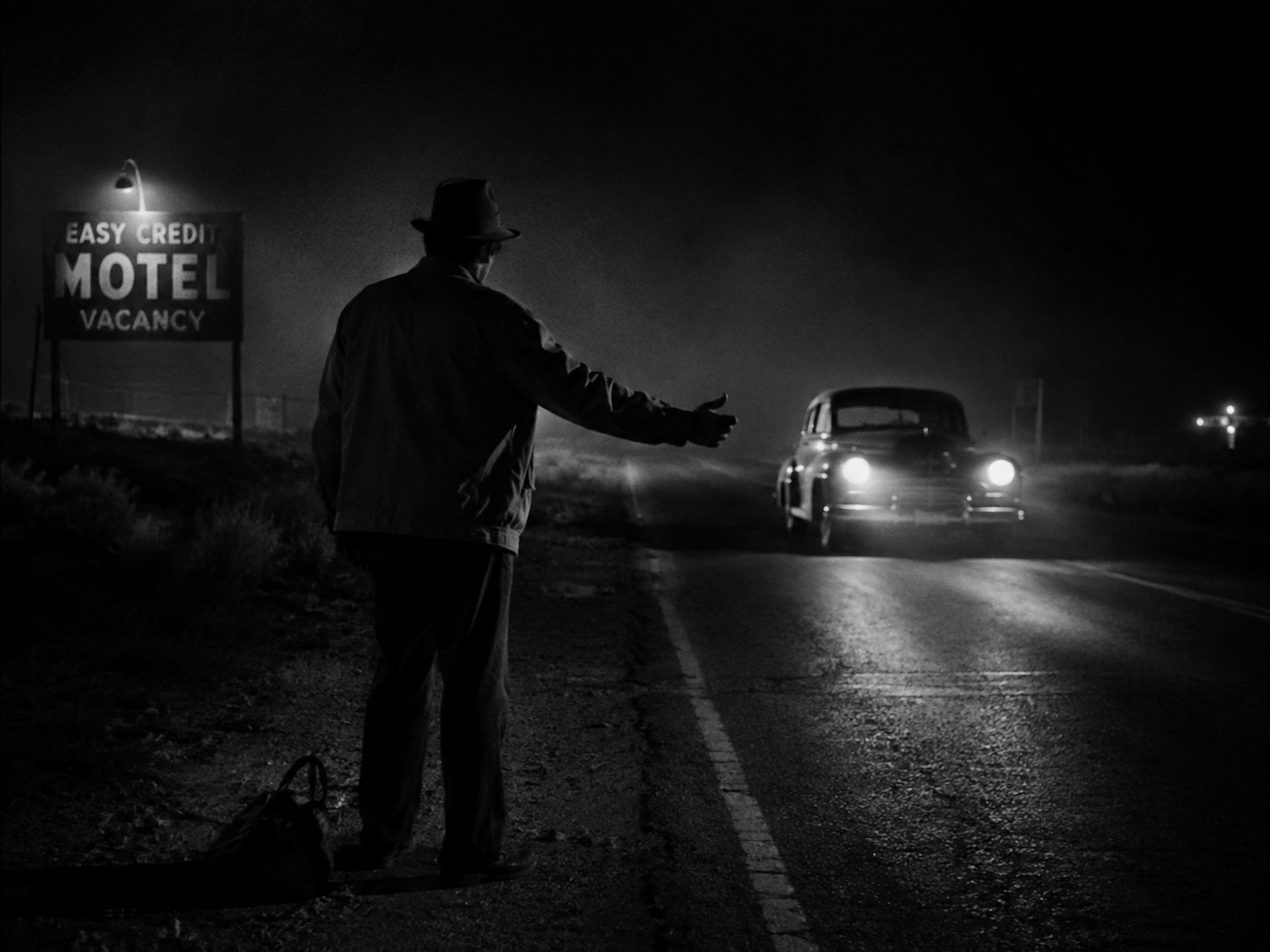 Film noir illustration of a lone hitchhiker near a motel on a dark highway