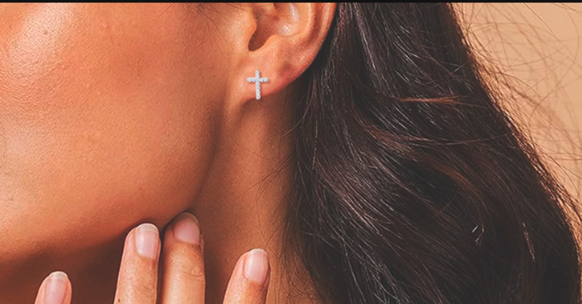 A close-up of a woman's neck, hand, and ear. She wears a cross stud earring and has long, brown hair.